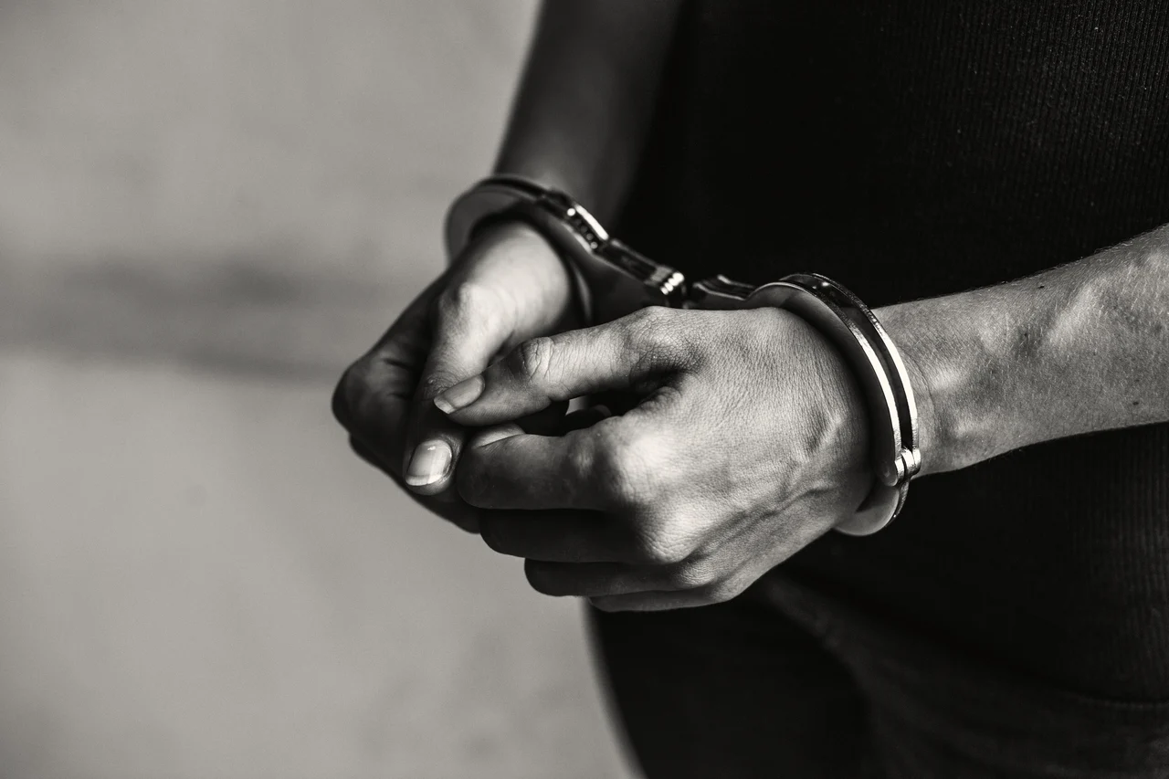 Homem aparece de costas, com as mãos algemadas, enquanto é acompanhado por um agente em uma rua de paralelepípedos.