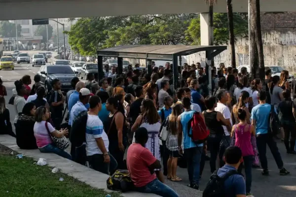 Ônibus municipal do Rio parado em ponto de embarque, com vários passageiros aguardando para entrar no veículo e trânsito ao fundo.