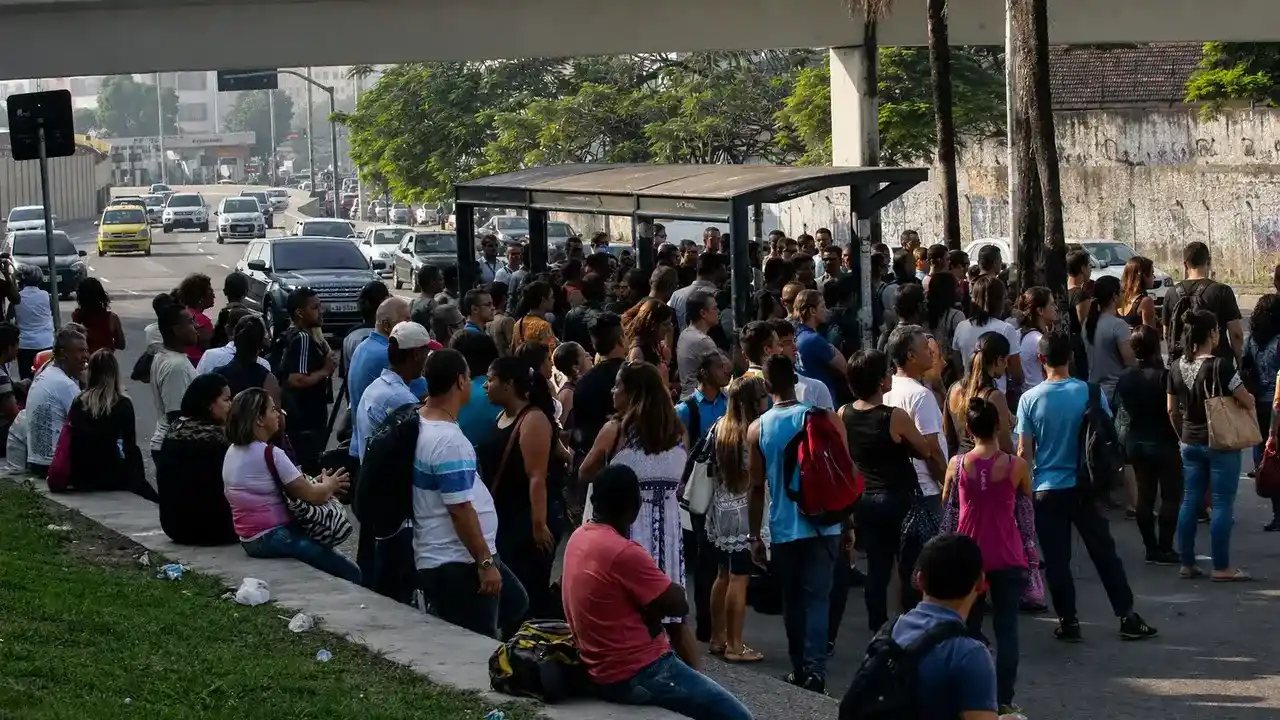 Ônibus municipal do Rio parado em ponto de embarque, com vários passageiros aguardando para entrar no veículo e trânsito ao fundo.