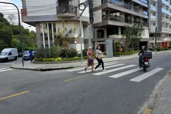 Duas mulheres atravessam cruzamento movimentado no Centro de Nova Friburgo enquanto veículos passam próximo a elas.