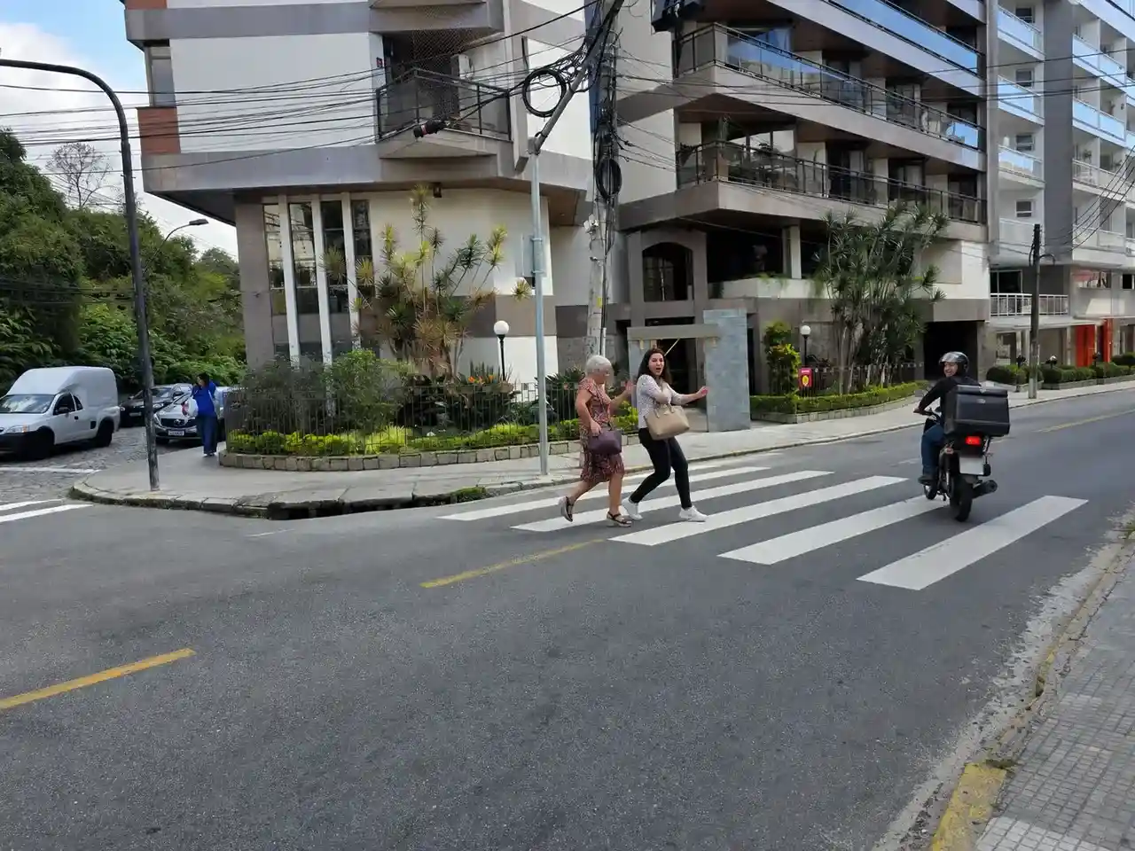 Duas mulheres atravessam cruzamento movimentado no Centro de Nova Friburgo enquanto veículos passam próximo a elas.