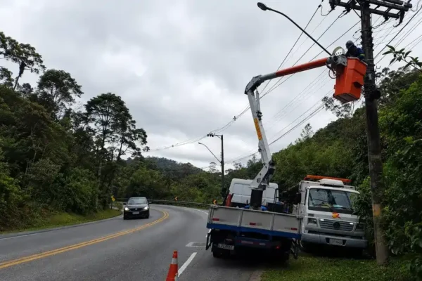 Funcionários realizam poda de árvores às margens da RJ-116 enquanto veículos passam pela rodovia sob operação Pare e Siga.