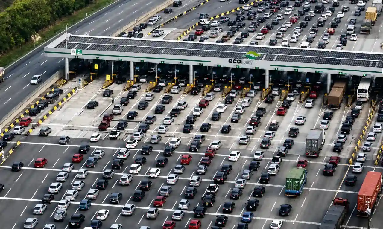A imagem mostra uma praça de pedágio em uma rodovia paulista, com várias cabines de cobrança e veículos passando pelas pistas.