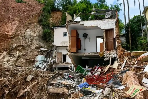 Imagem mostra uma casa parcialmente destruída por um deslizamento de terra. A estrutura ficou aberta após o desabamento, com móveis ainda visíveis entre os escombros e o barranco ao fundo.