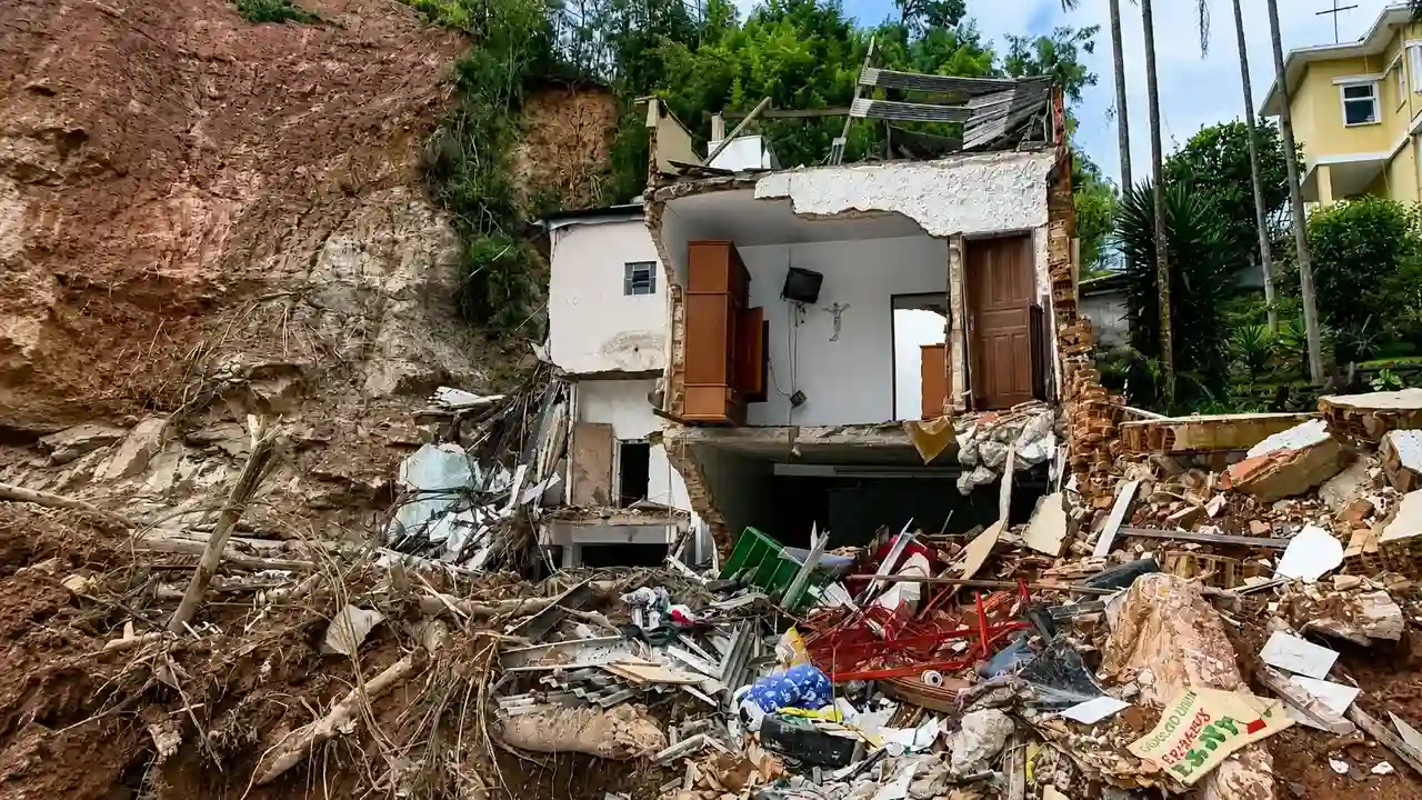 Imagem mostra uma casa parcialmente destruída por um deslizamento de terra. A estrutura ficou aberta após o desabamento, com móveis ainda visíveis entre os escombros e o barranco ao fundo.