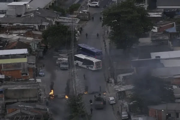 Imagem aérea mostra ônibus usados para bloquear uma rua em Costa Barros, com focos de incêndio, fumaça e presença policial na região.