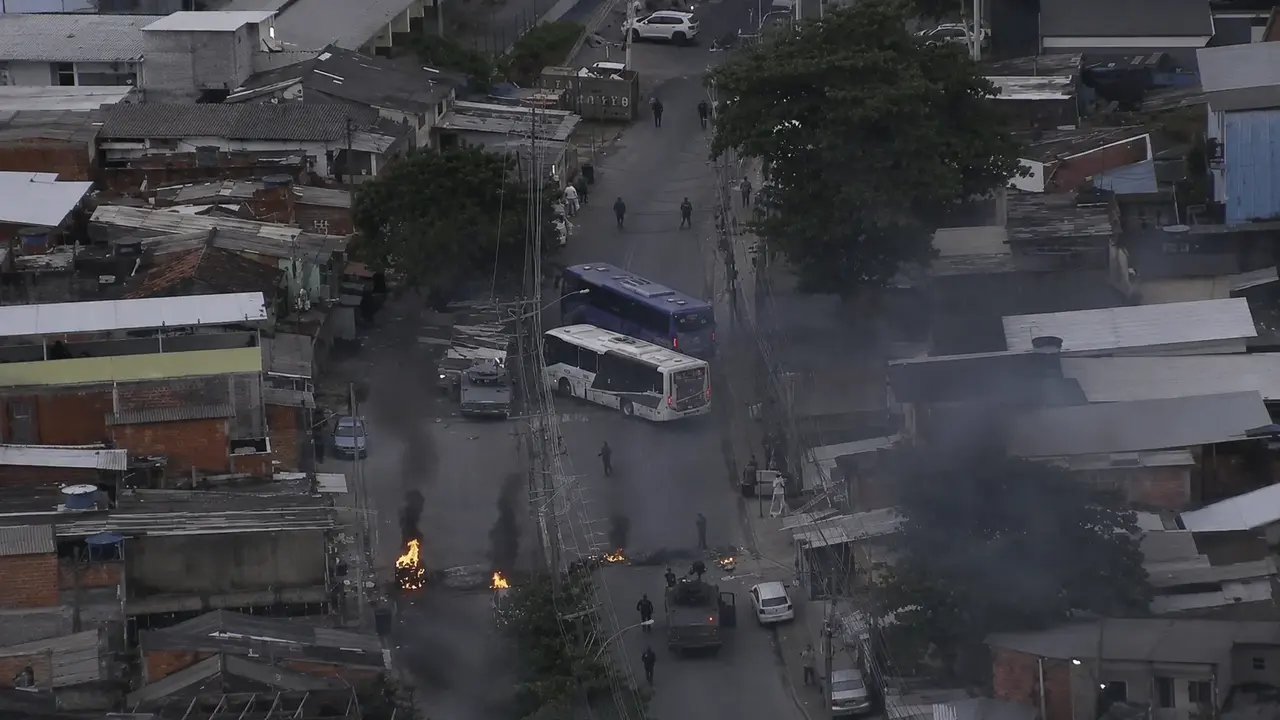Imagem aérea mostra ônibus usados para bloquear uma rua em Costa Barros, com focos de incêndio, fumaça e presença policial na região.