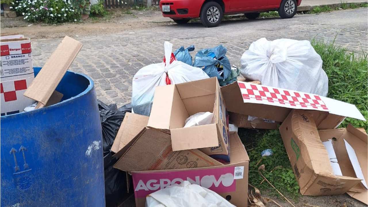Acúmulo de lixo na rua, com caixas de papelão, sacos plásticos e um tambor azul.