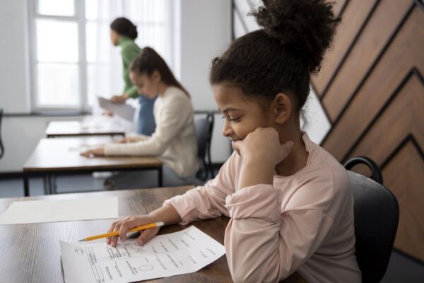 Aluna concentrada em prova escolar, com colegas e professora ao fundo.