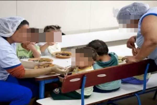 Auxiliares de creche servindo crianças em refeitório, com rostos censurados.