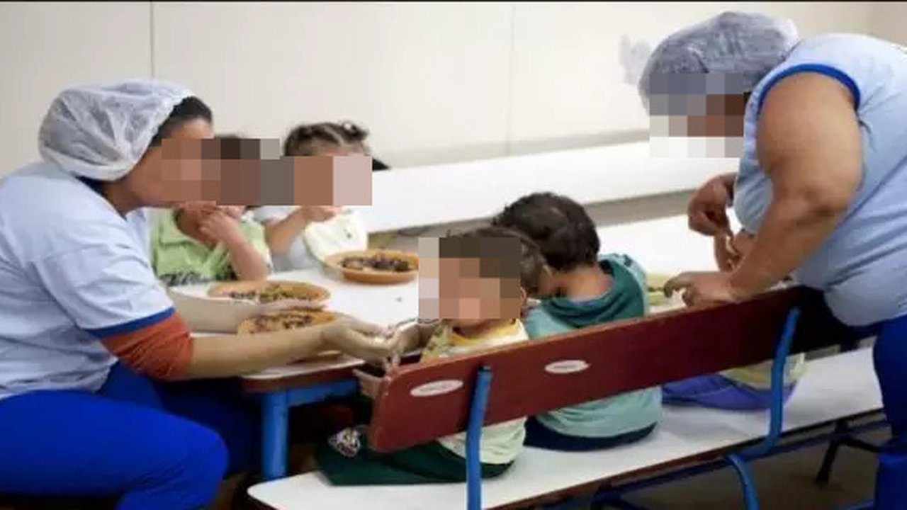 Auxiliares de creche servindo crianças em refeitório, com rostos censurados.
