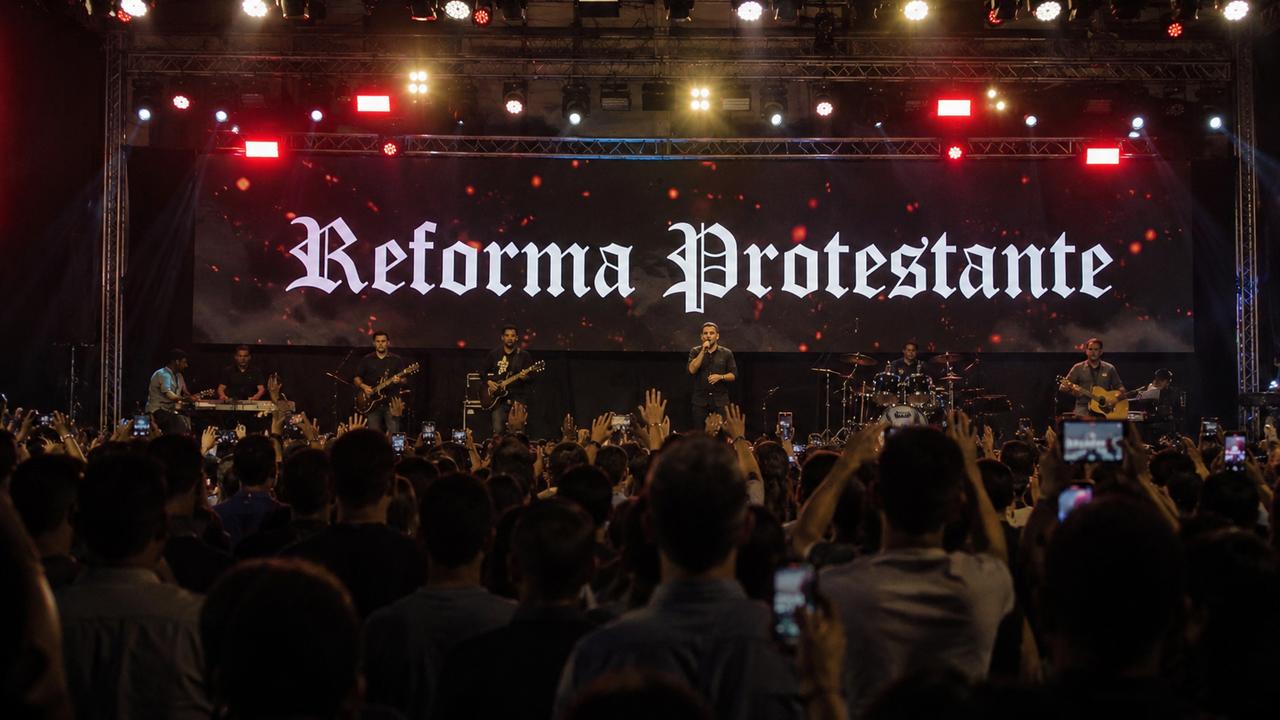 Banda se apresentando no evento Reforma Protestante com a palavra 'Reforma Protestante' no telão.