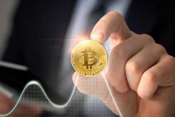 Bitcoin sendo segurado por uma mão, com gráfico de análise financeira ao fundo, representando valorização.