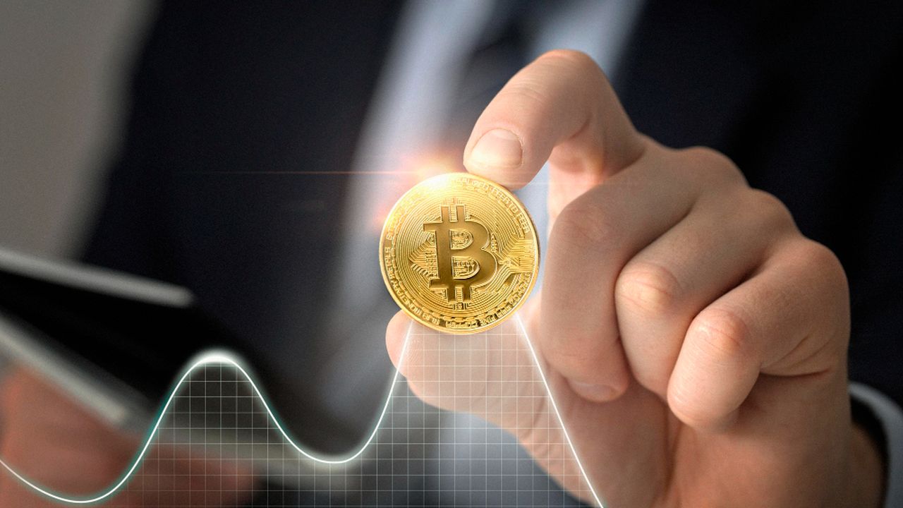 Bitcoin sendo segurado por uma mão, com gráfico de análise financeira ao fundo, representando valorização.