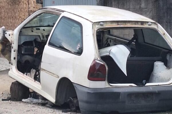 Carcaça de carro branco em estado de abandono, sem rodas e com partes removidas.