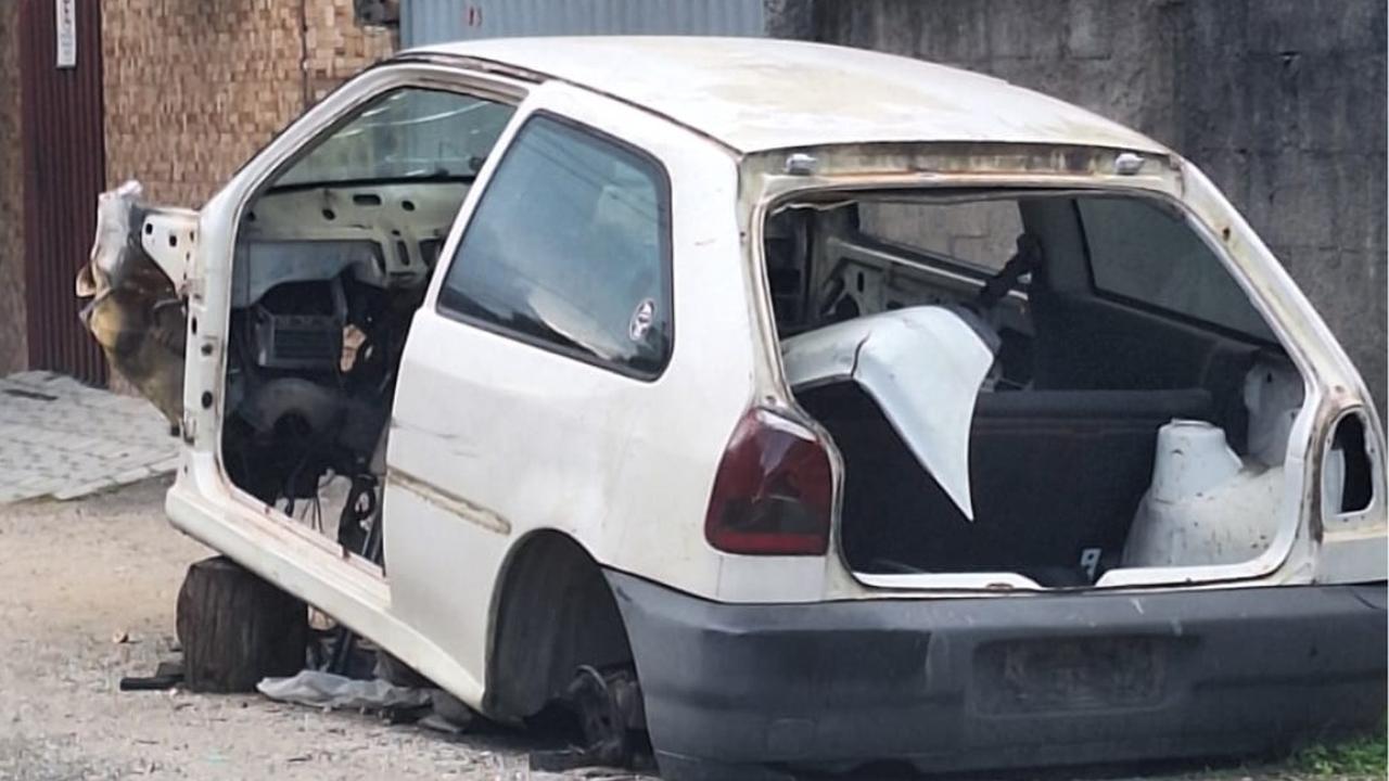 Carcaça de carro branco em estado de abandono, sem rodas e com partes removidas.