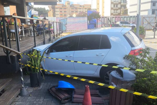 Carro branco danificado em estacionamento de estabelecimento comercial, com faixa de isolamento.
