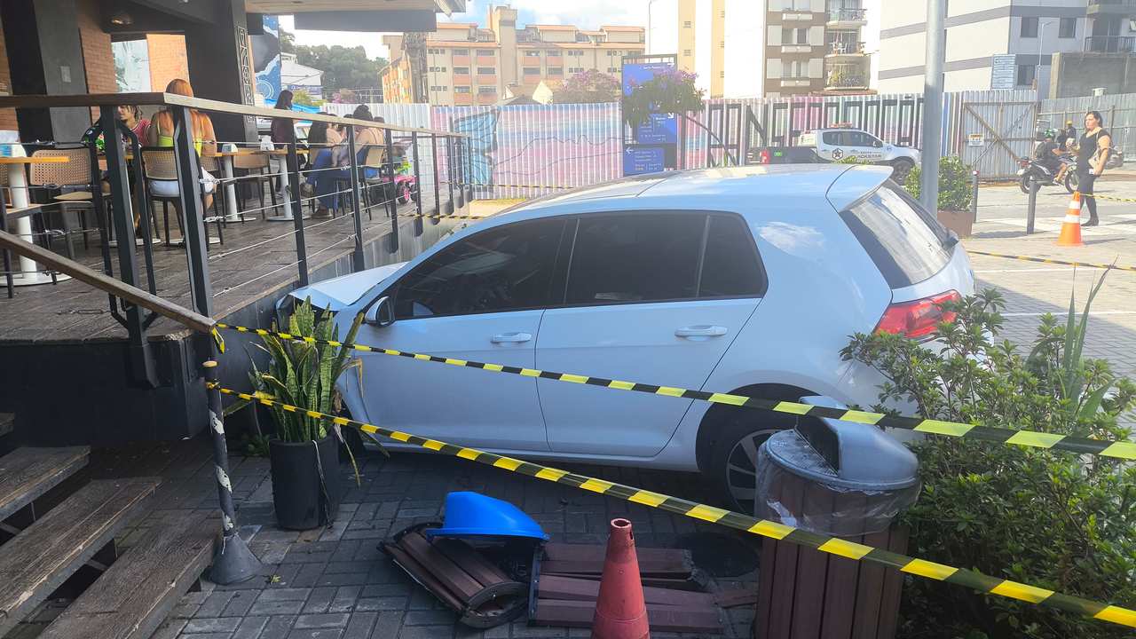 Carro branco danificado em estacionamento de estabelecimento comercial, com faixa de isolamento.