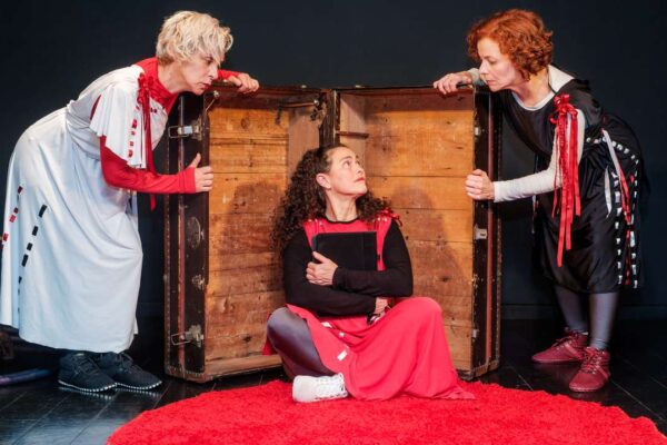 Cena teatral com três atrizes interagindo perto de um baú antigo.