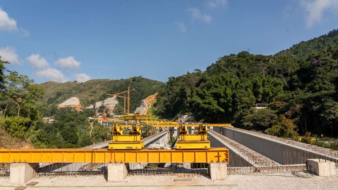 Construção de ponte em região montanhosa com equipamentos e natureza ao fundo.
