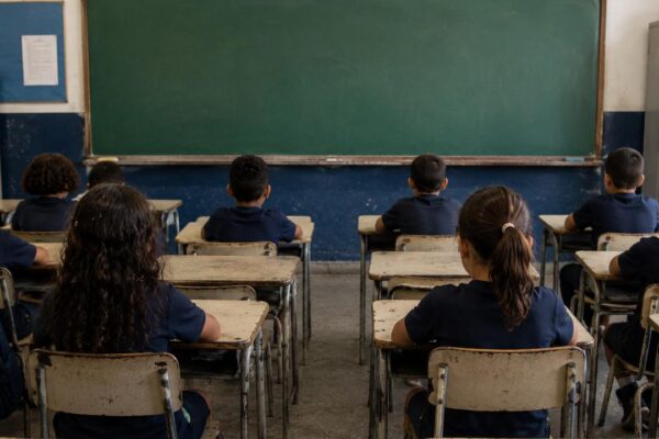Crianças em sala de aula, com foco em suas costas, sentadas em carteiras escolares.