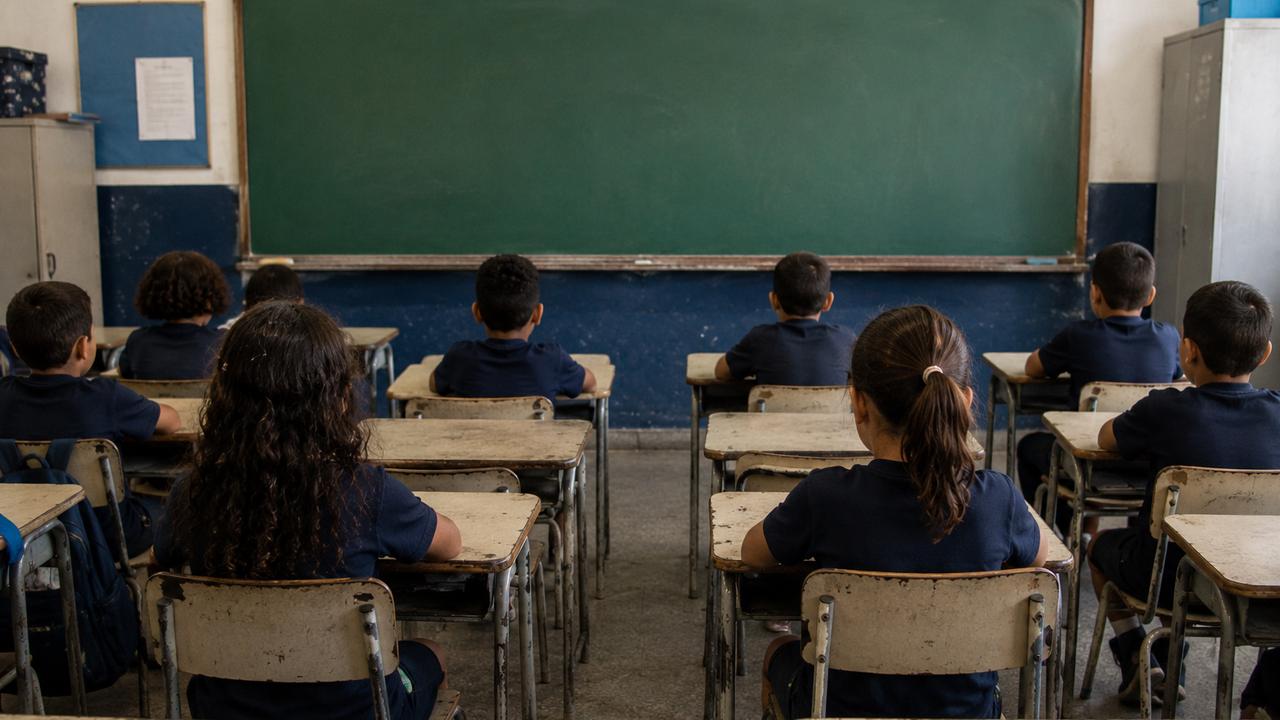 Crianças em sala de aula, com foco em suas costas, sentadas em carteiras escolares.