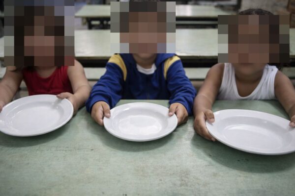 Crianças seguram pratos vazios em mesa, com rostos pixelados, em contexto de escassez alimentar.