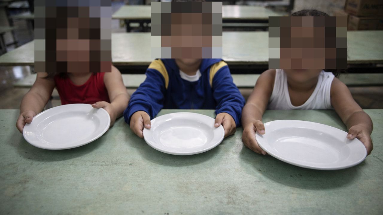Crianças seguram pratos vazios em mesa, com rostos pixelados, em contexto de escassez alimentar.
