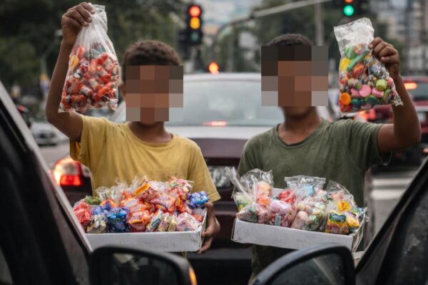 Crianças vendendo doces em rua, em meio a carros e trânsito, com rostos pixelados.