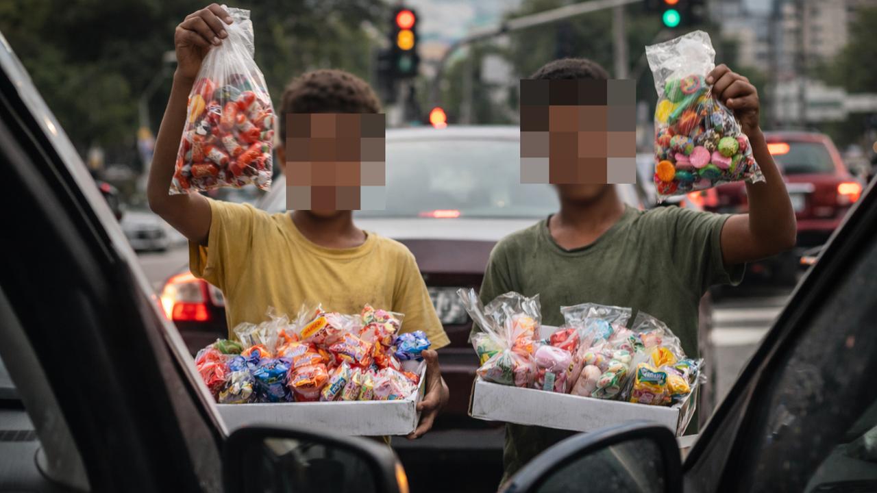 Crianças vendendo doces em rua, em meio a carros e trânsito, com rostos pixelados.