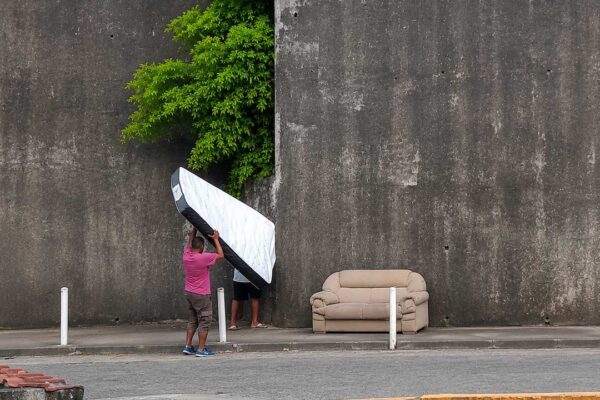 Dois homens carregam um colchão ao lado de um sofá abandonado em uma rua.