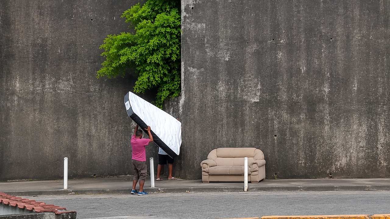 Dois homens carregam um colchão ao lado de um sofá abandonado em uma rua.