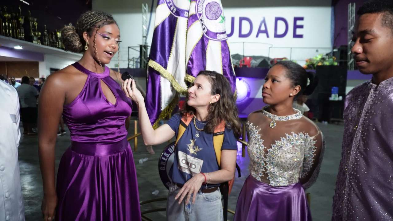 Entrevista com participantes do desfile de escolas de samba, com pessoas vestidas em trajes elaborados.
