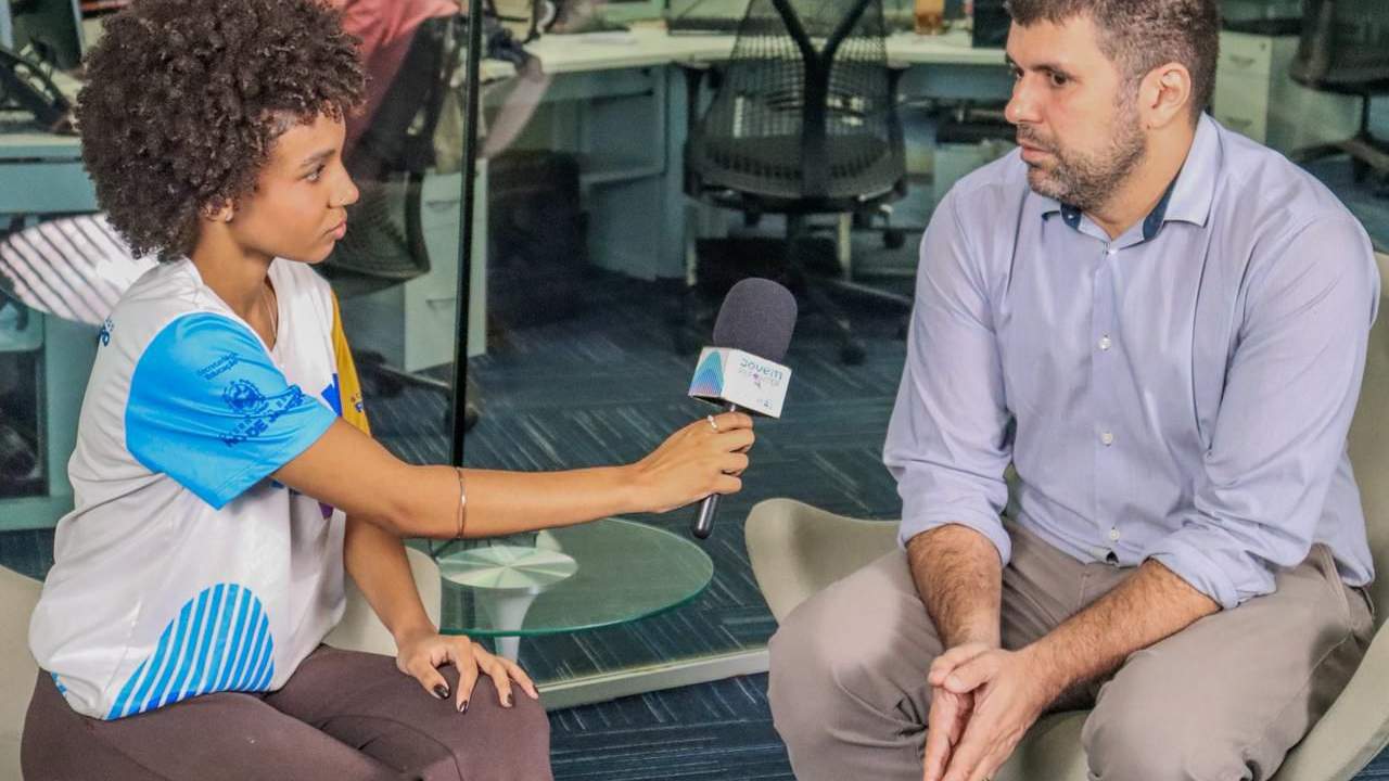 Entrevistadora com microfone entrevista homem em ambiente de escritório.
