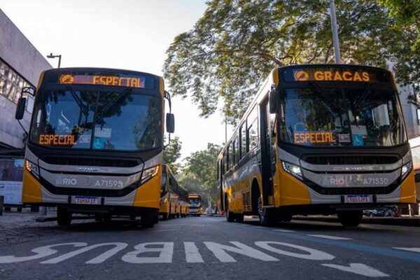 Dois ônibus novos do Sistema RIO estacionados em uma via urbana, com outros veículos ao fundo, durante apresentação da nova frota municipal.
