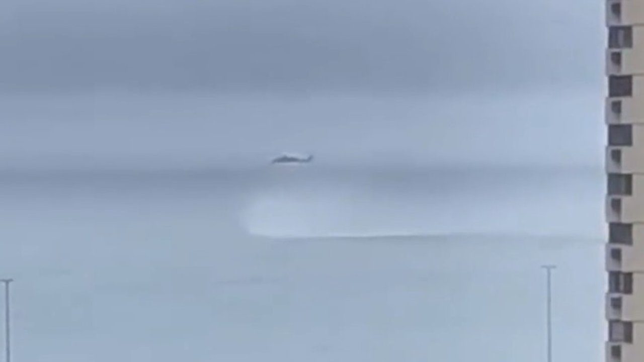 Helicóptero voando baixo sobre a água, com edifícios ao fundo.