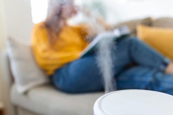 Humidificador em primeiro plano com pessoa lendo no sofá, ressaltando a importância da umidade do ar.