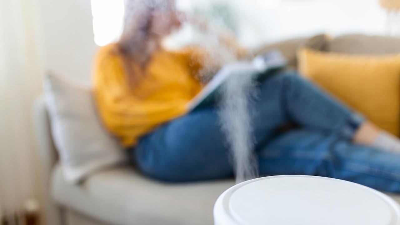 Humidificador em primeiro plano com pessoa lendo no sofá, ressaltando a importância da umidade do ar.