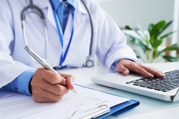 Médico escrevendo em prontuário e usando laptop, em contexto de investigação.