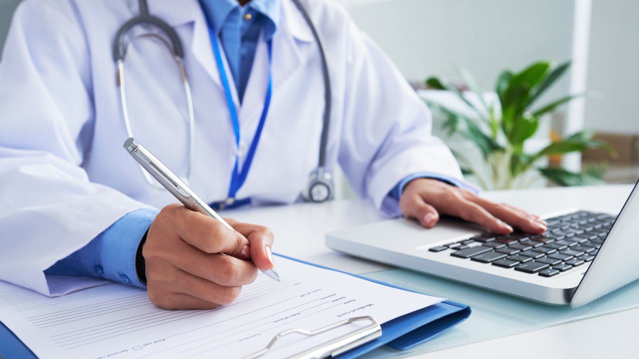 Médico escrevendo em prontuário e usando laptop, em contexto de investigação.
