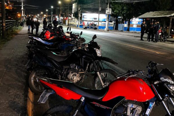 Motocicletas estacionadas em via pública à noite, com pessoas e polícia ao fundo