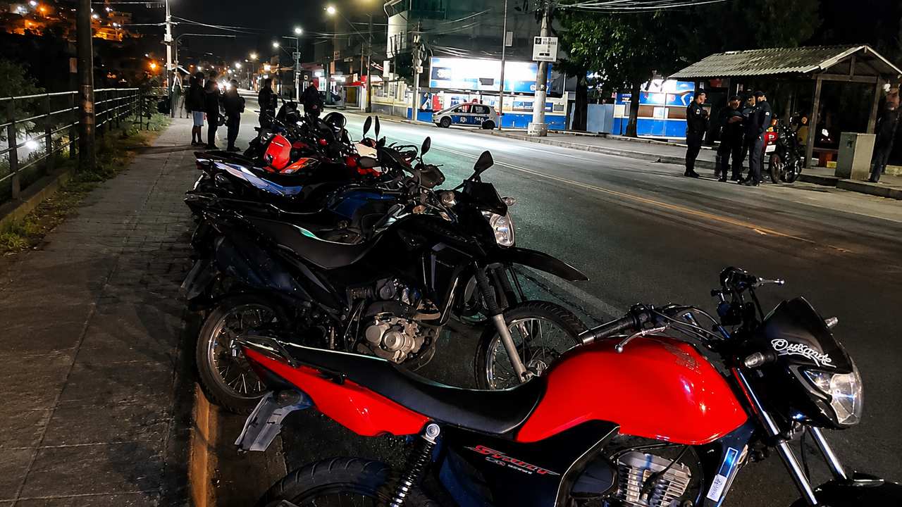 Motocicletas estacionadas em via pública à noite, com pessoas e polícia ao fundo