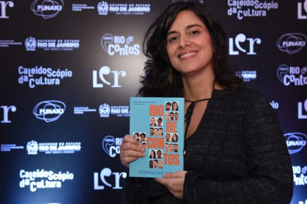 Mulher sorrindo segura livro 'Rio de Contos' em evento literário.