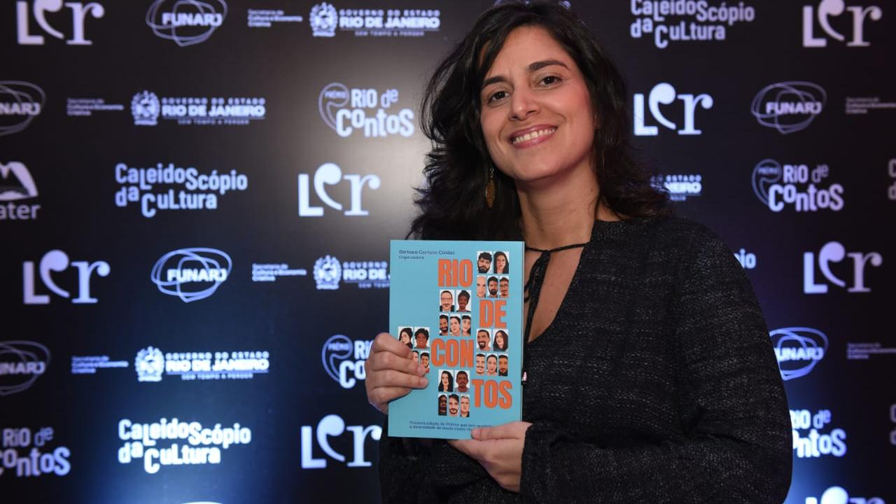Mulher sorrindo segura livro 'Rio de Contos' em evento literário.