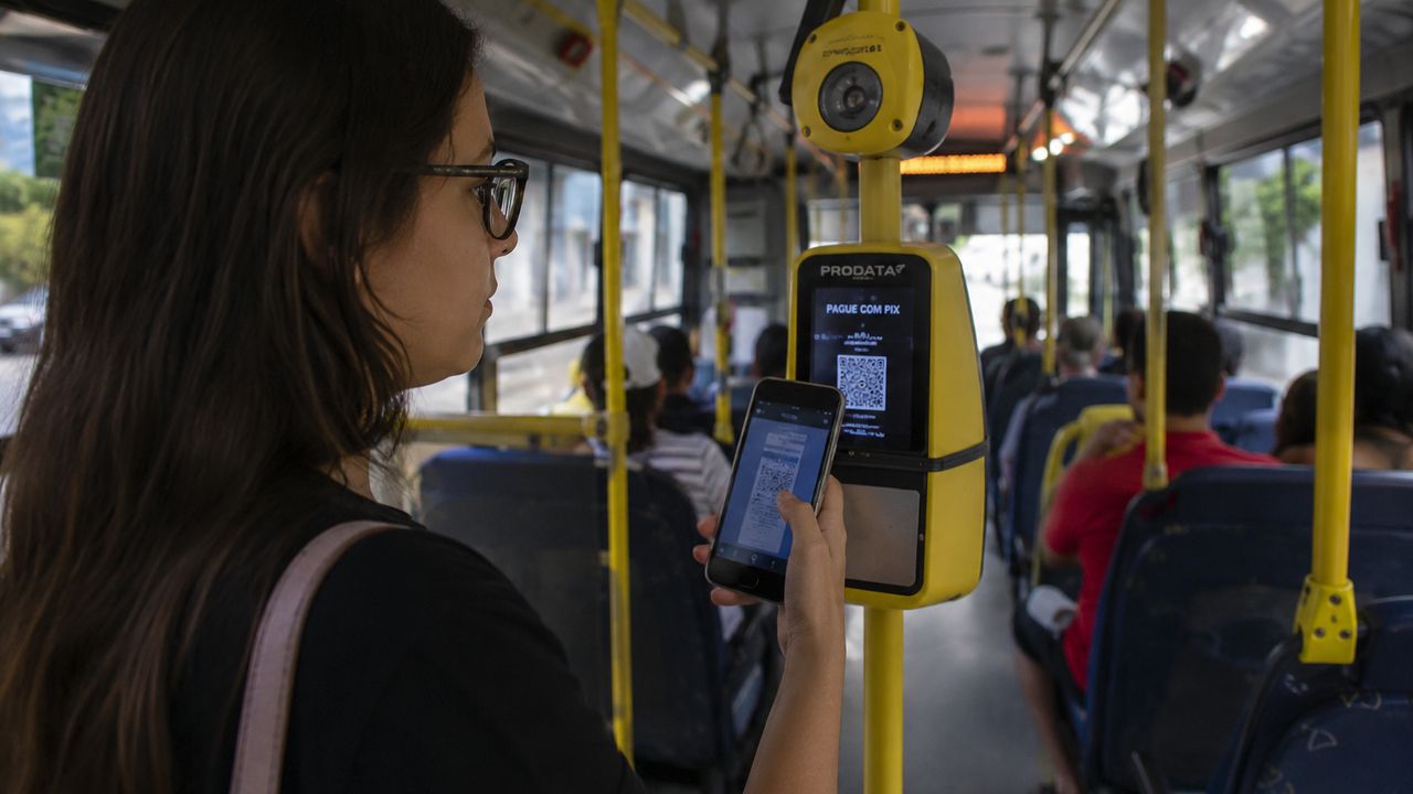 Mulher utilizando aplicativo para pagar passagem de ônibus com celular, no Brasil.