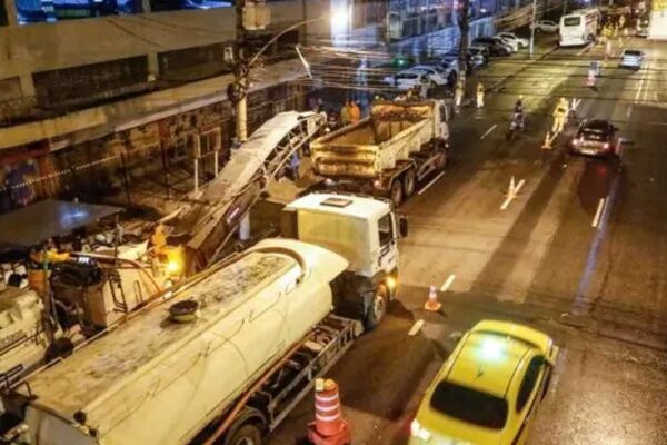 Obras de recapeamento noturno em via urbana, com máquinas e caminhões.