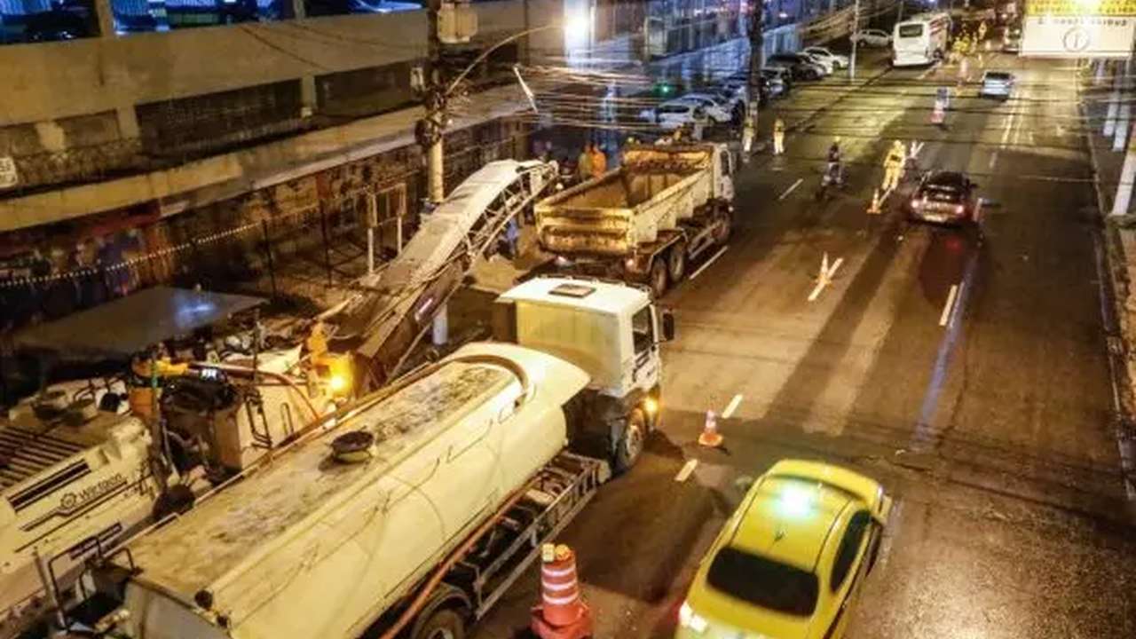 Obras de recapeamento noturno em via urbana, com máquinas e caminhões.