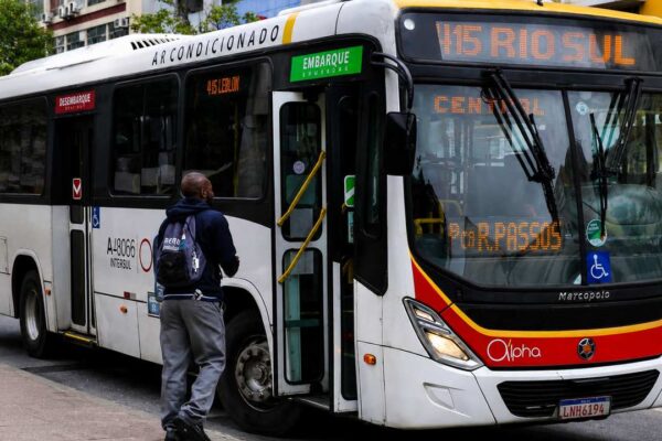 Ônibus branco com detalhes em vermelho e amarelo, com um passageiro embarcando.