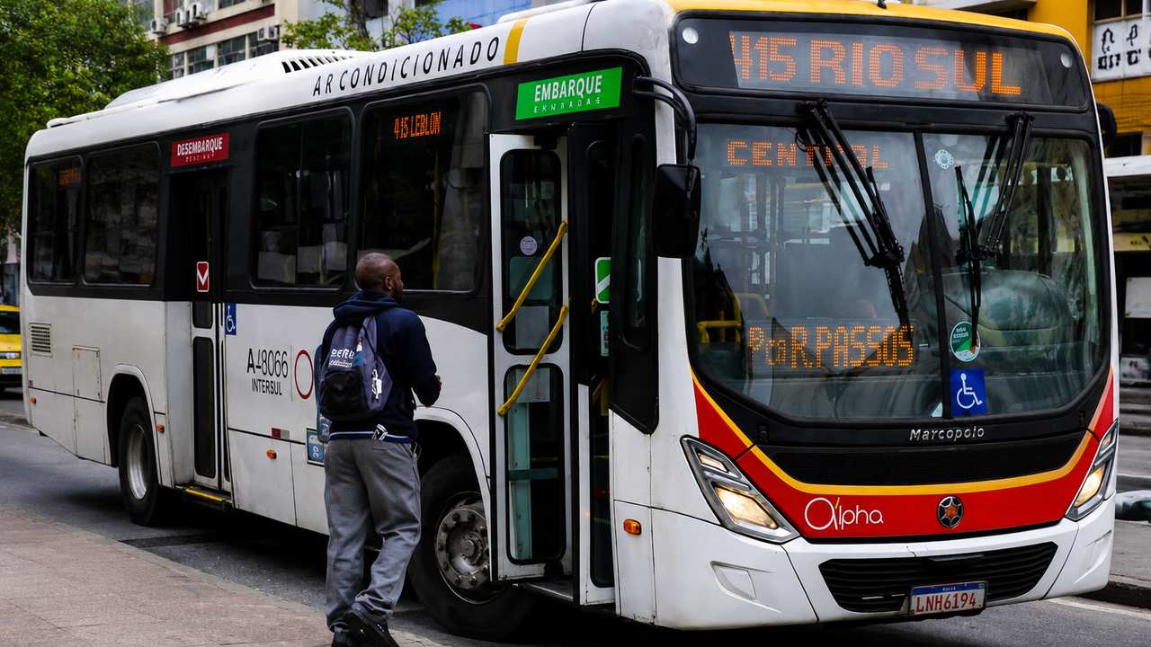 Ônibus branco com detalhes em vermelho e amarelo, com um passageiro embarcando.