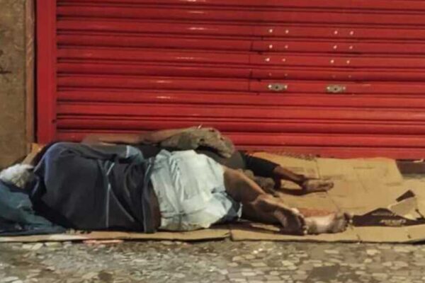 Pessoas dormindo na rua em frente a uma estrutura comercial fechada, com sinalização grafitada.